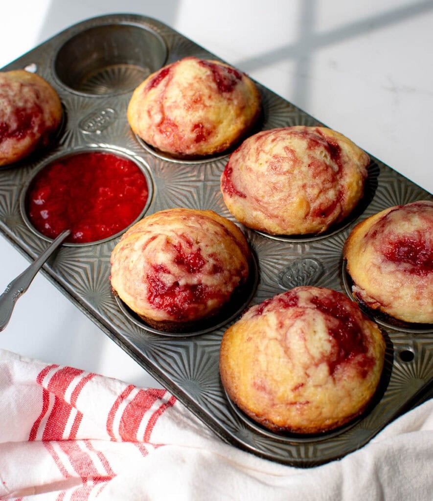 A pan of freshly baked strawberry muffins with one of the muffin tins filled wth strawberry sauce.