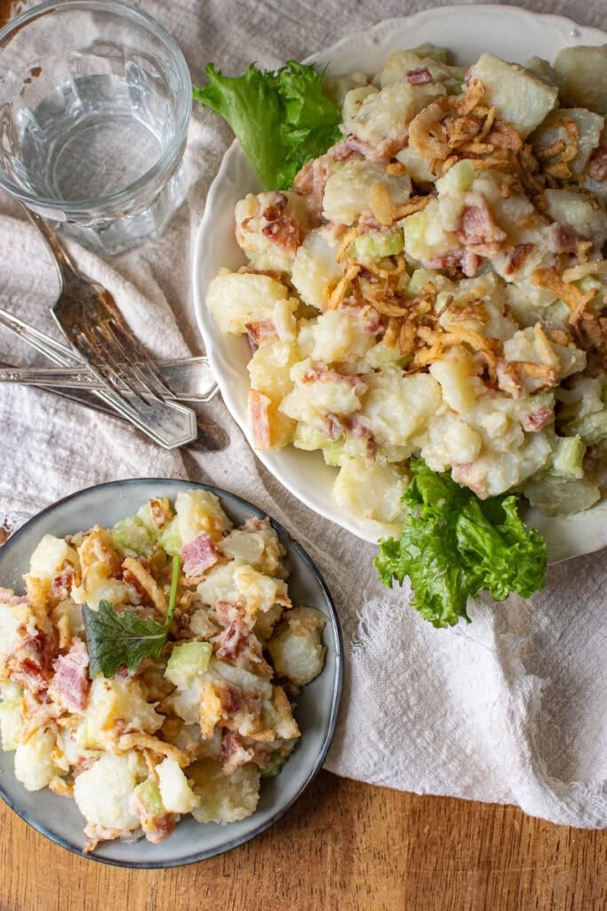 A large bowl of potato bacon salad garnished with greens, a small plate with a serving of the salad along side a fork and glass of water.
