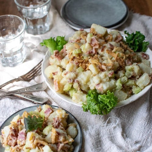 Potato and bacon salad in a serving bowl with a serving on a small plate, 2 empty plates waiting to be filled and 2 water glasses to the side.