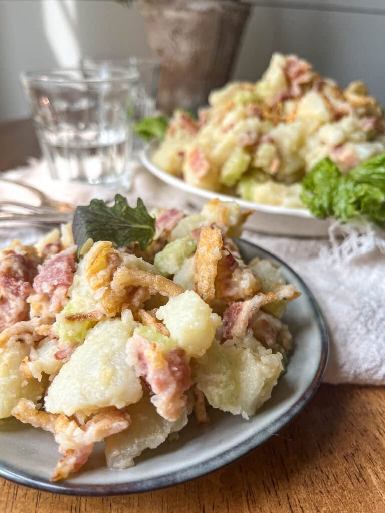 A small serving a potato bacon salad in the foreground with a full serving bowl full in the background.