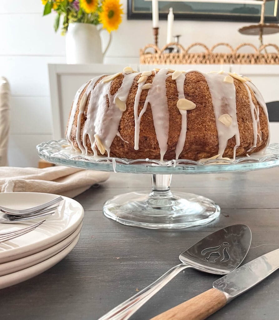 Zucchini bundt cake on a clear cake plate, drizzled with almond glaze and a sprinkle of sliced almonds.