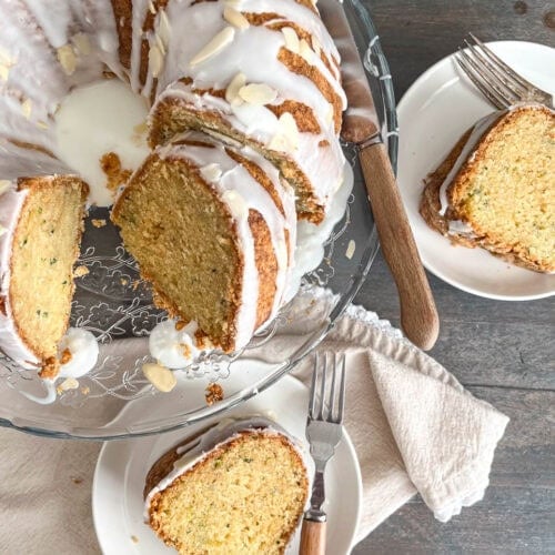A zucchini bundt cake with almond glaze and slivered almonds, with one slice cut out and on a white plate.