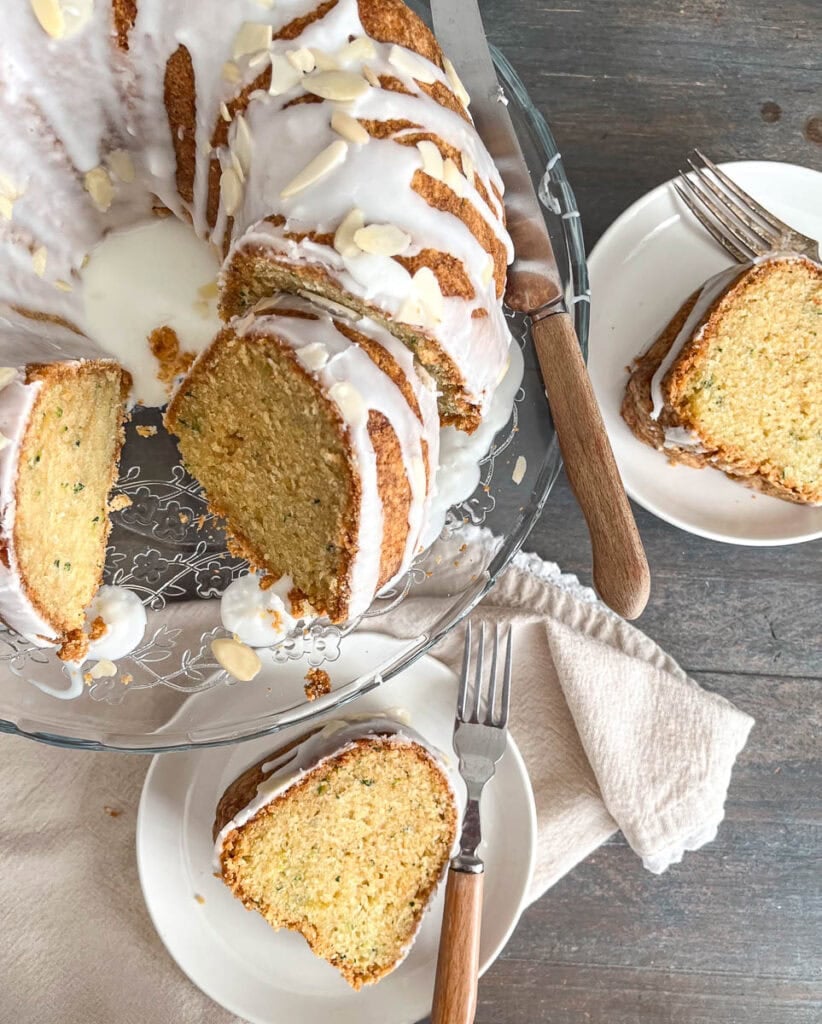 A zucchini bundt cake with almond glaze and slivered almonds, with one slice cut out and on a white plate.