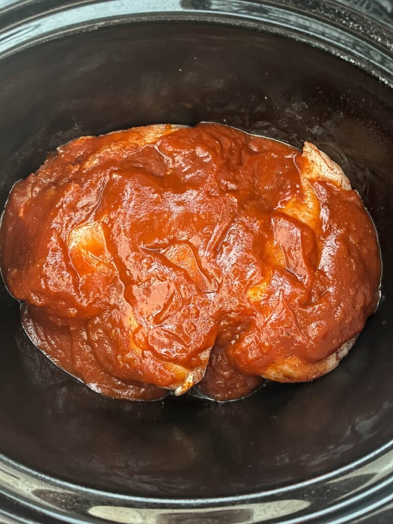 Seasoned chicken breast covered in barbecue sauce in the slow cooker before cooking.