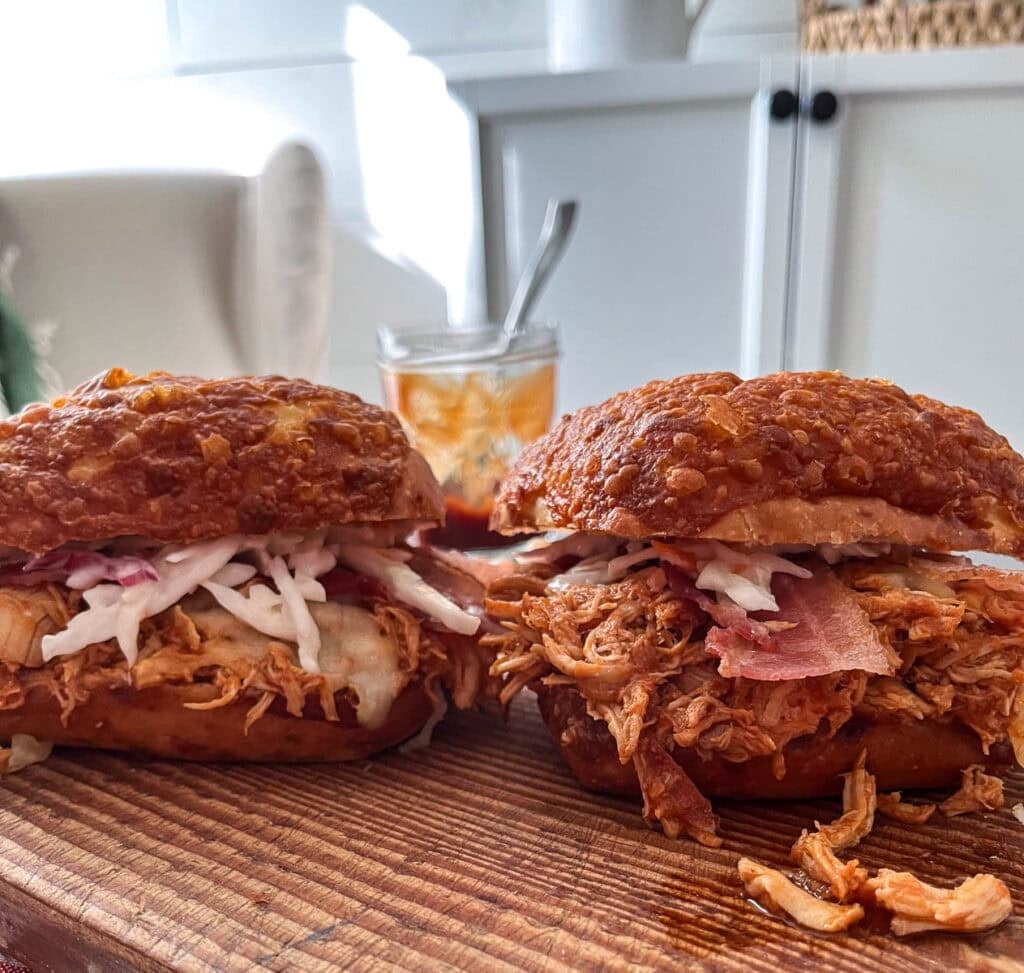 Two pulled chicken sandwiches topped with melted cheese, bacon and coleslaw on a cutting board.