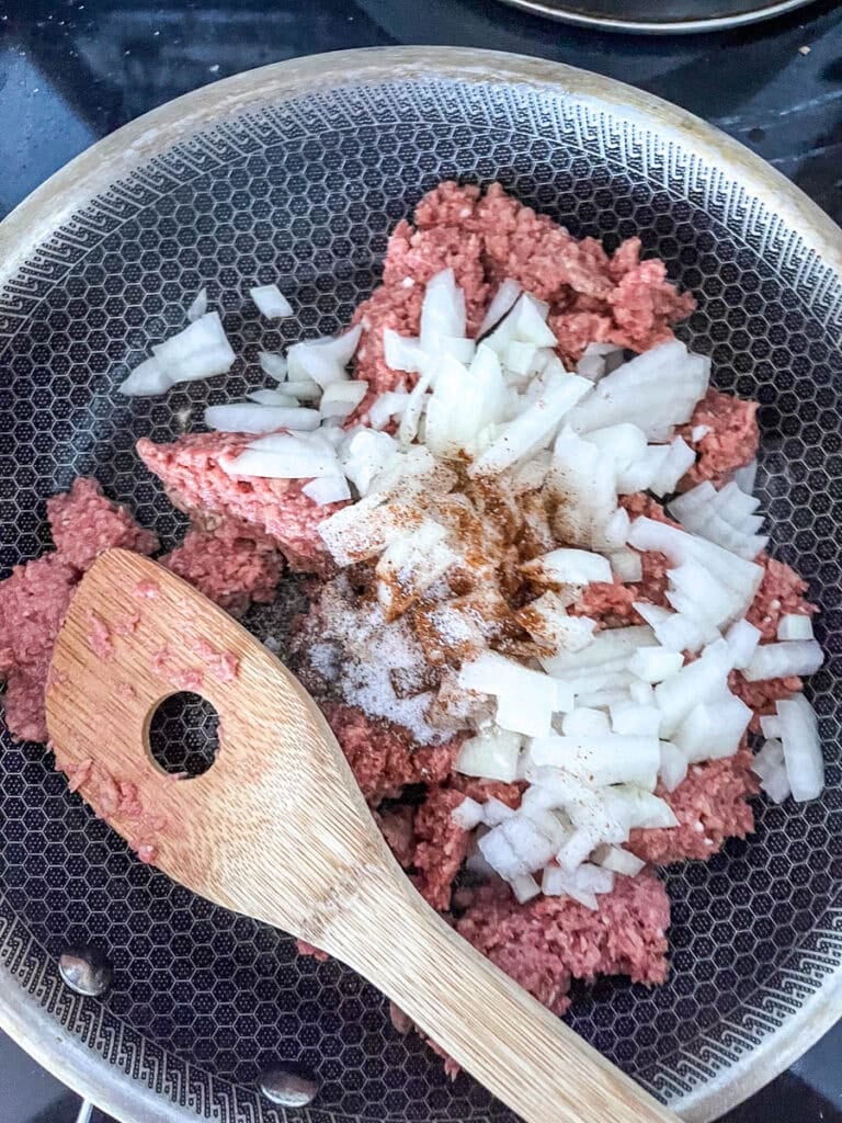 A pound of ground beef, chopped onions and chilli powder ready to cook in a medium frying pan.