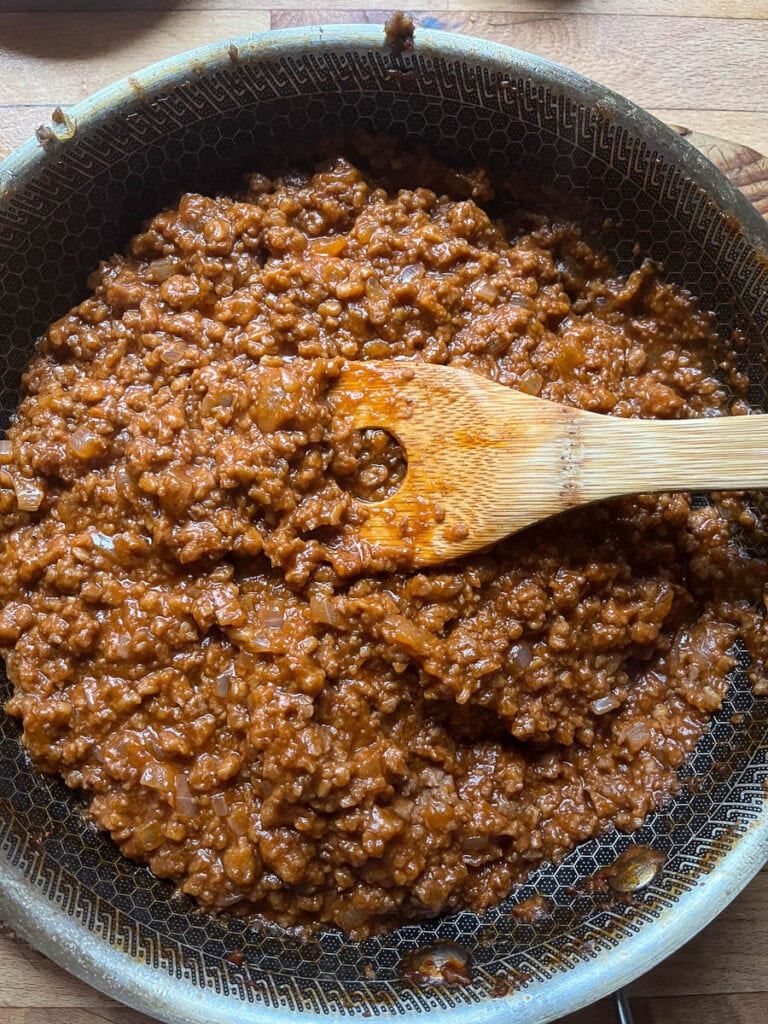 Fully cooked sloppy Joe filling in a frying pan with a wooden spoon in the pan.