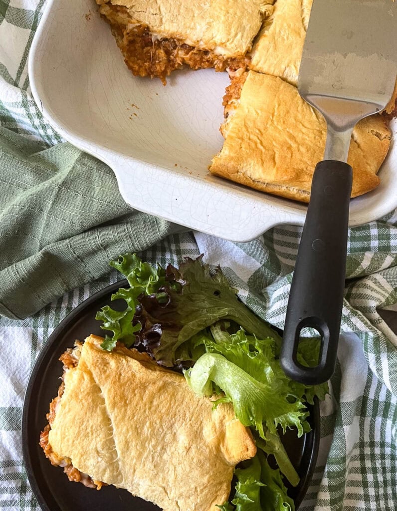 Sloppy Joe casserole in a white casserole dish with one slice cut out and placed on a black dinner plate with a side salad.