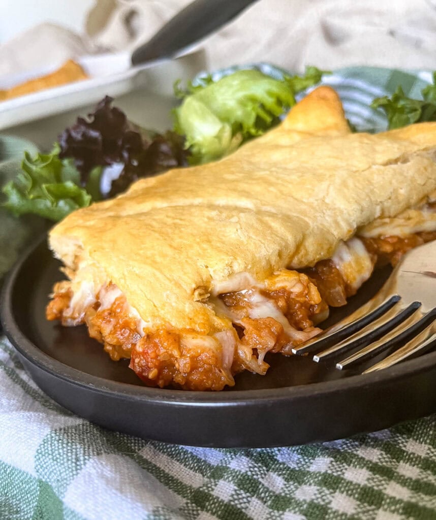 A slice of crescent roll sloppy Joe casserole on a black dinner plate with a side salad, and a dinner fork.