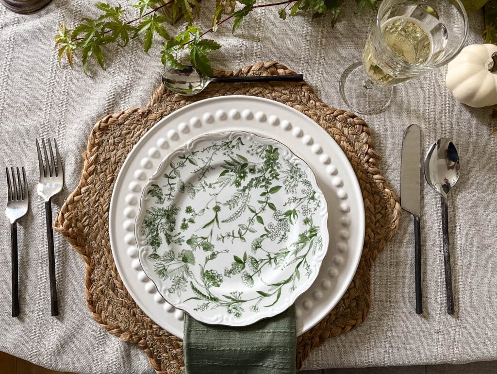 Overhead view of an English Cottage place setting with twig handled flatware, woven placemat, simple footed wine glass, white dinner plate and green botanical salad plate.