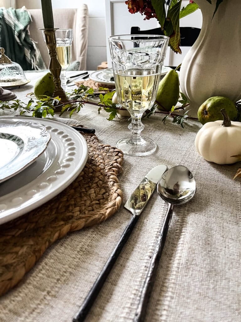 An English Cottage place setting with twig handled flatware, woven placemat, simple footed wine glass, white dinner plate and green botanical salad plate.