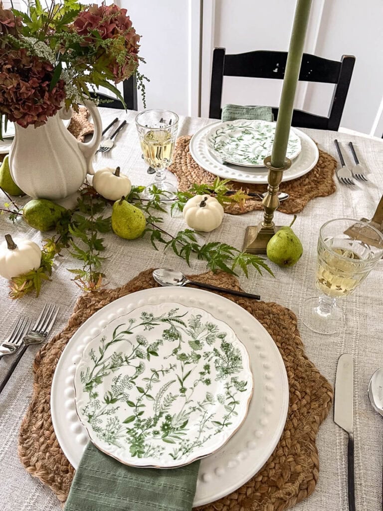 Two place settings on a table set for autumn that features woven place mats with scalloped edges, white dinner plates topped with green botanical lunch plates, fresh miniature Japanese maple branches, fresh pears and faux cream coloured pumpkins.