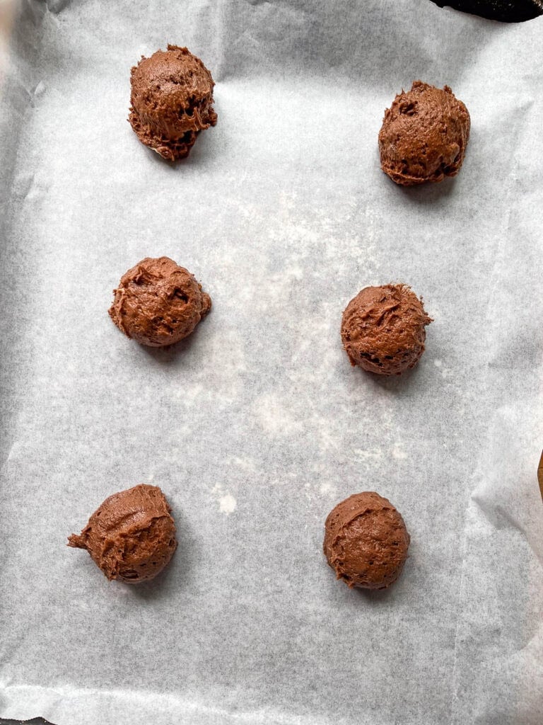 Six large scoops of brownie cookie batter on a parchment lined baking sheet before baking.