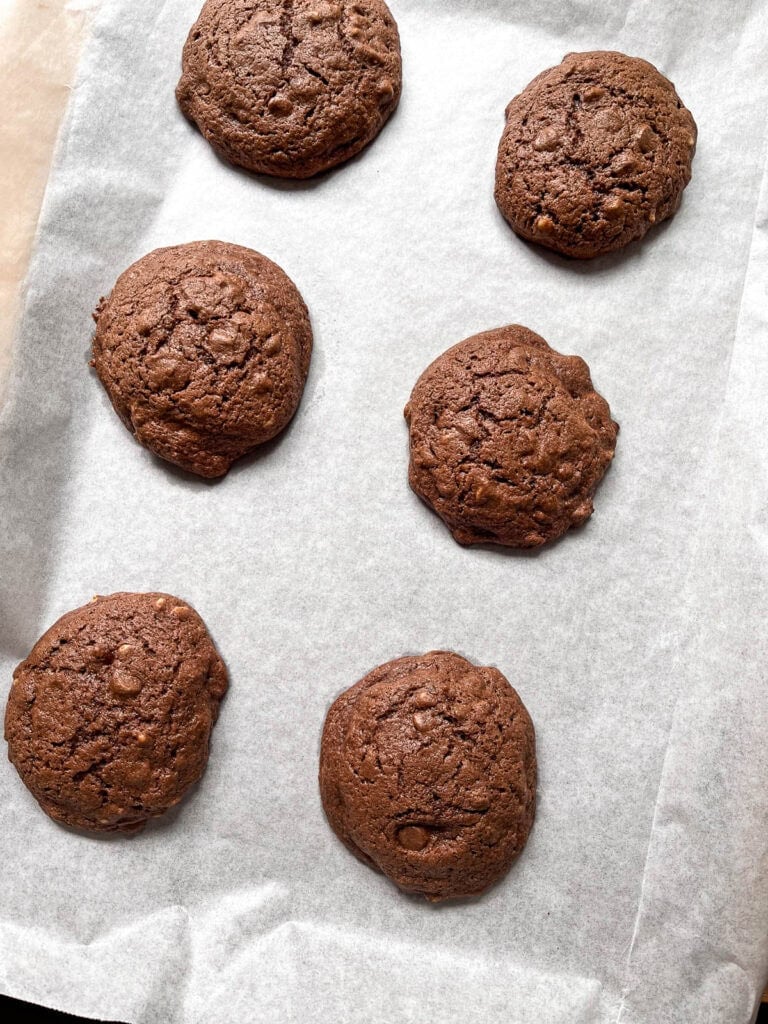 Six large freshly baked chocolate chip brownie cookies on a parchment lined baking sheet.