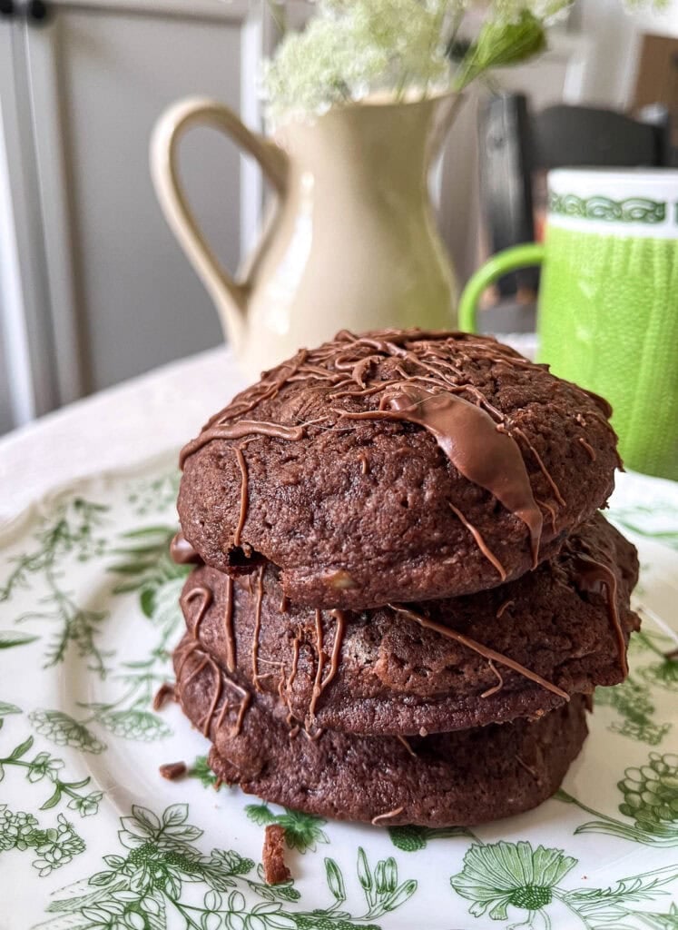 A stack of 3 brownie cookies with chocolate drizzle on a white plate with a green botanical print.