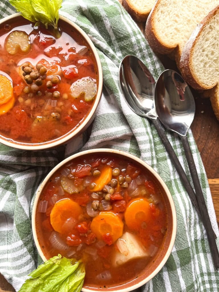 Two bowls of Autumn Soup made with carrots, potatoes, diced tomatoes and lentils alongside 2 soup spoons and slices of bread, all on a white and green tea towel.