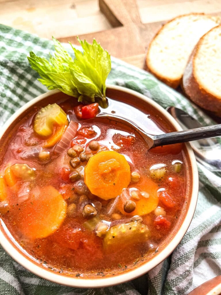 A close up of a bowl of Autumn soup with carrots, sliced celery, diced tomatoes, potatoes and lentils, garnished with a celery greens and slices of bread on the side.
