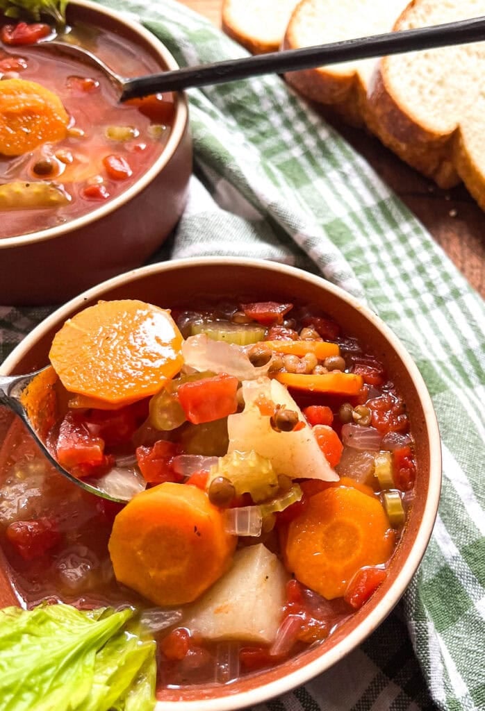 A bowl of a tomato based vegetable soup in a brown bowl, with a spoon full of soup lifted out of the bowl.