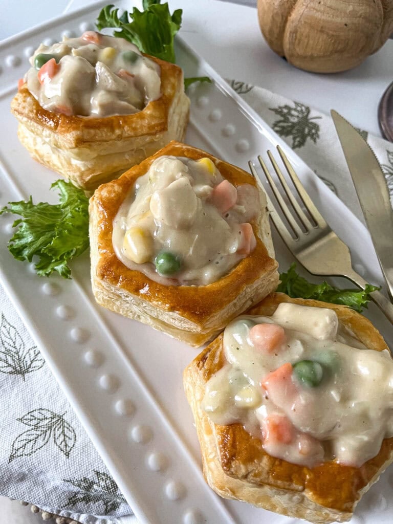 Three puff pastry shells filled with chicken pot pie filling on a long platter, and garnished with lettuce leaves.