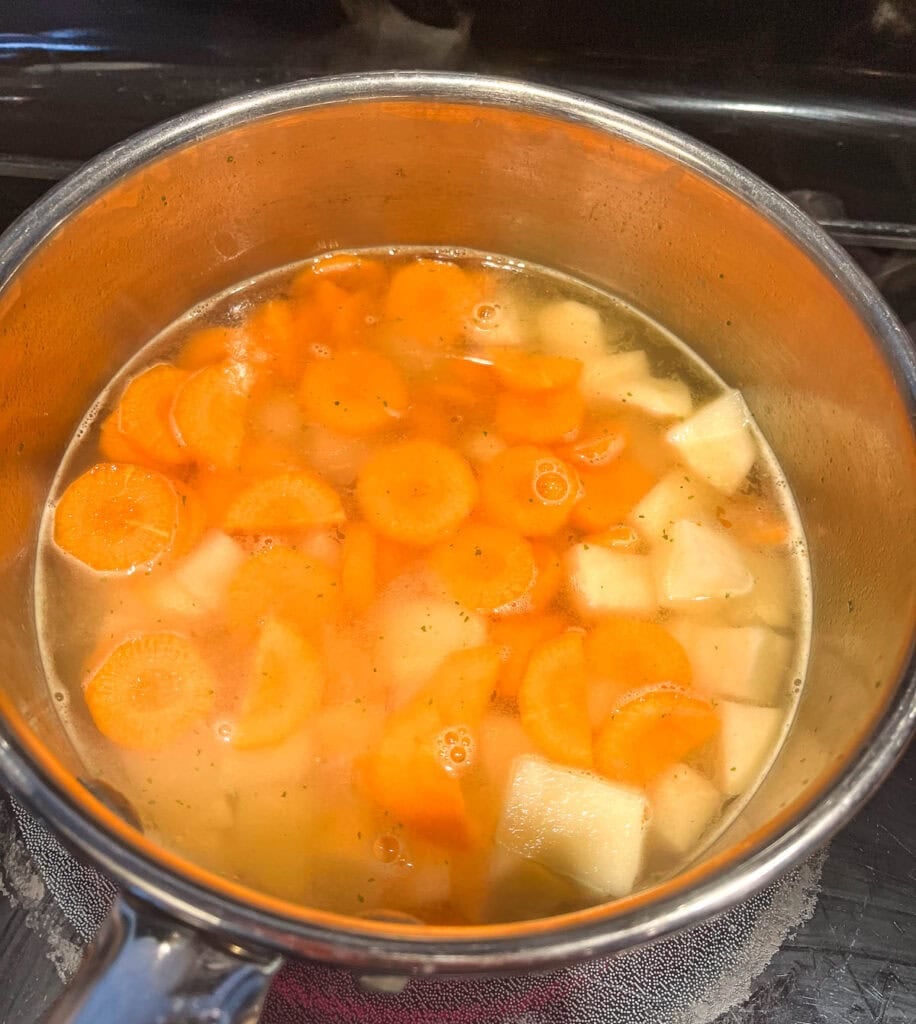 Carrots and cubed potatoes cooking in a base of water and chicken bouillon in a saucepan on the stove top.