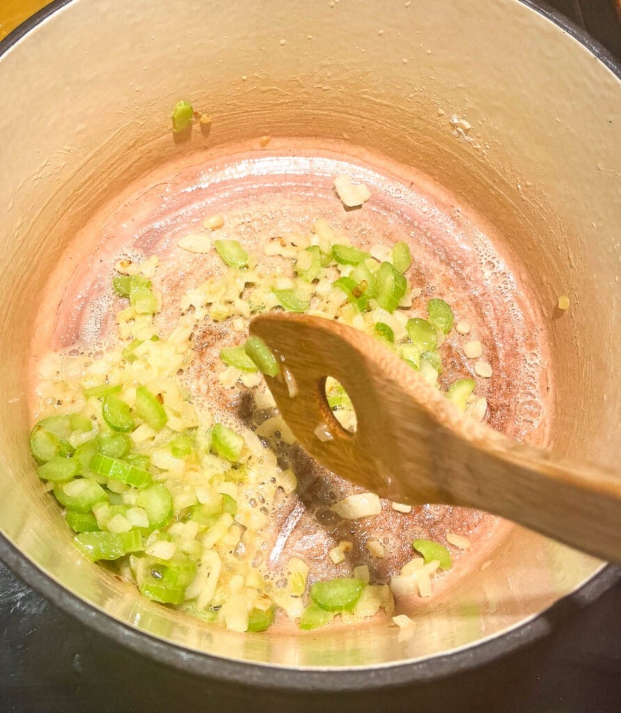 Flour added to softened onions and celery in a dutch oven on the stove top, being stirred with a wooden spoon to cook out the raw flour flavour and create a roux. 