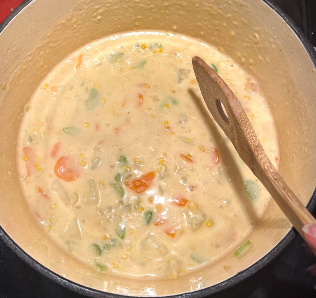 Cooked potatoes and carrots and a can of creamed corn added to the creamy chowder base to create corn and potato chowder simmering on the stove top in a dutch oven.