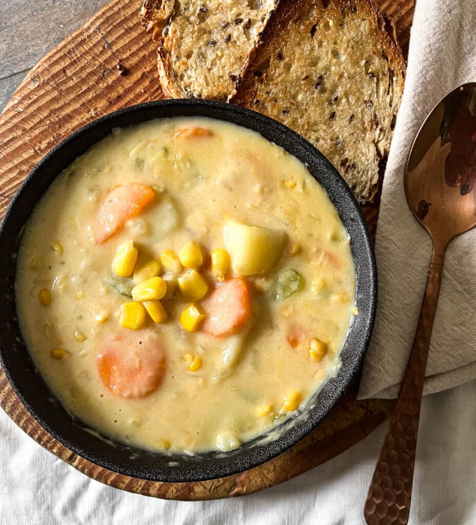 A bowl of corn and potato chowder with 2 slices of toast on a cutting board and a napkin and spoon off to the side.