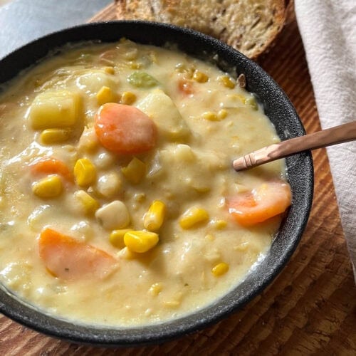 A bowl of potato corn chowder on a cutting board with a copper spoon in the bowl.