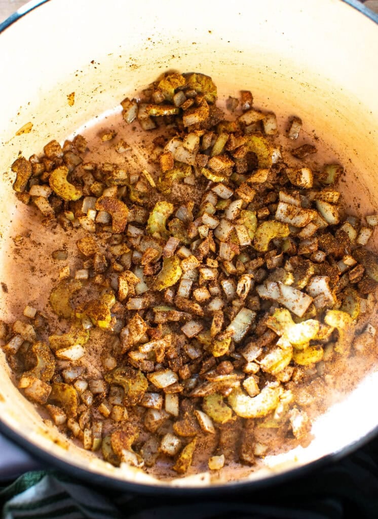 Diced onions and celery that have been cooked until soft, stirred together with chili spices in a dutch oven on the stove top.