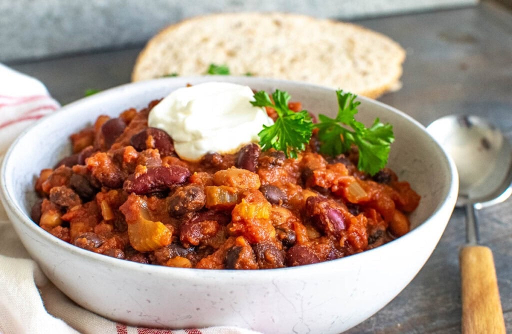 A bowl of 3 bean chili garnished with a dollop of sour cream and fresh parsley, and a slice of bread on the side.
