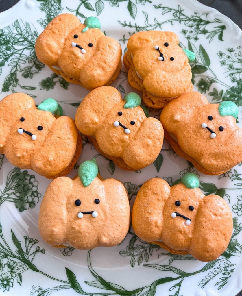 A plate of orange macarons in the shaped of pumpkins with little faces drawn on.