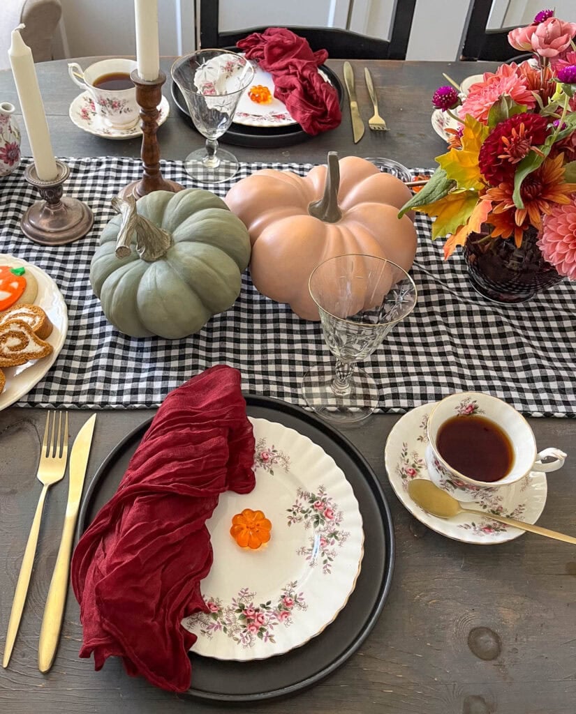 A table set for afternoon tea at Halloween with green and orange pumpkins, fine china with pink and burgundy flowers and dark burgundy napkins.