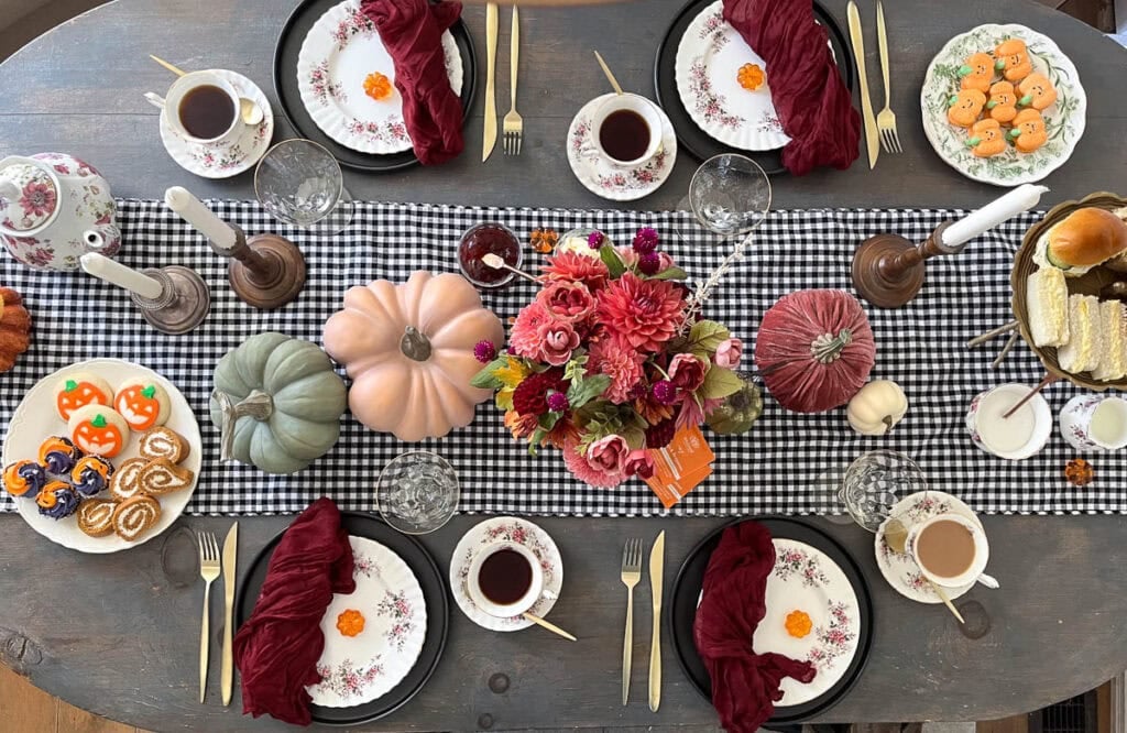 An overhead view of a table styled for a Halloween afternoon tea with a black and white check table runner, moody coloured floral arrangement, white floral plates, and various pumpkins.