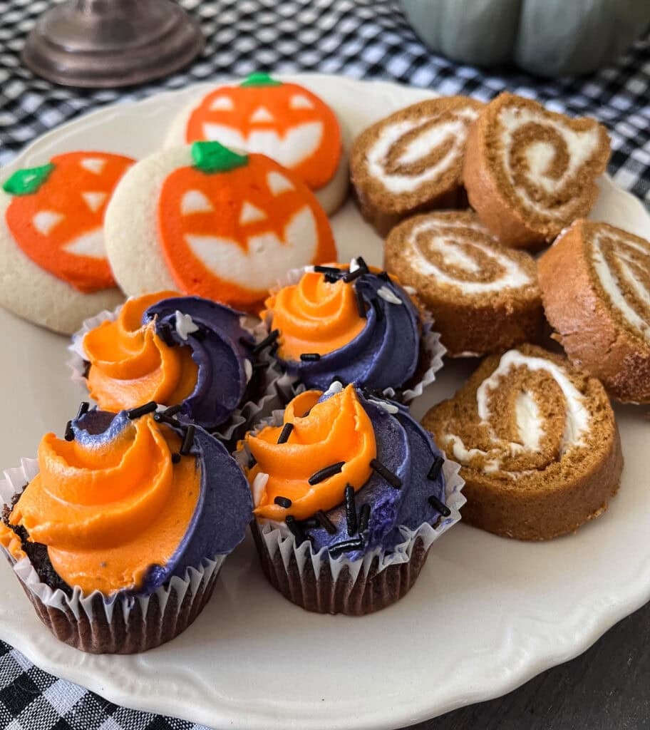 A plate of Halloween sweet treats including jack-o-lantern soft sugar cookies, orange and purple decorated mini cupcakes, and slices of pumpkin spice roll cake.