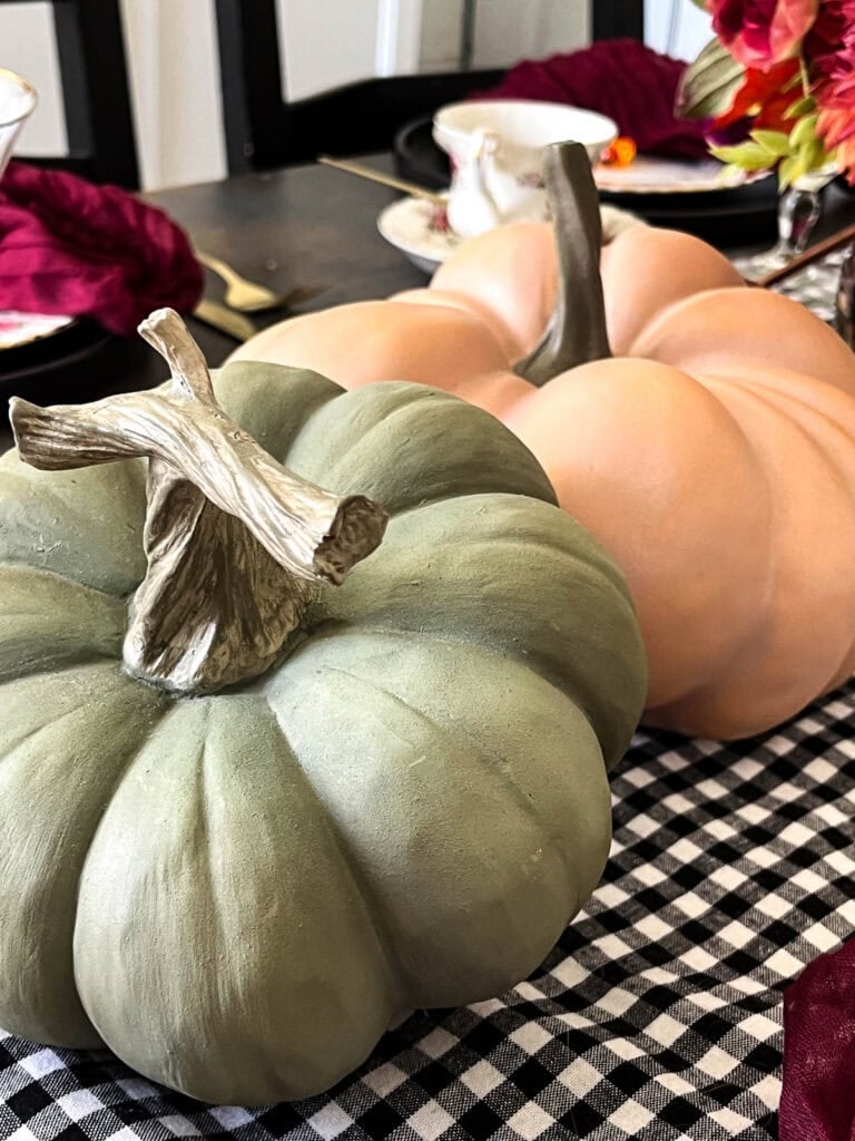 Two large ceramic pumpkins arranged on a dining table. One is soft orange in colour and the other sage green.